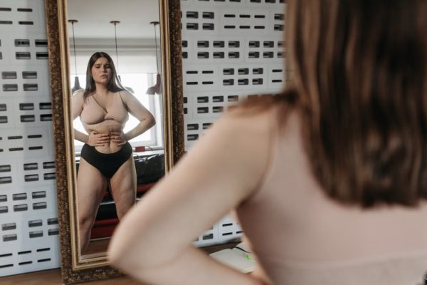 Woman in underwear looking at her reflection in a mirror, contemplating body image.