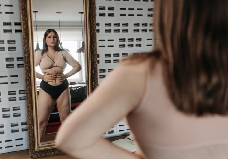Woman in underwear looking at her reflection in a mirror, contemplating body image.